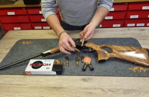 Hände reinigen eine Repetierbüchse auf einer Werkbank, daneben liegt Munition